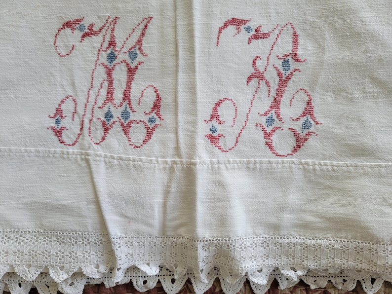May include: Cream-colored linen textile with embroidered red and blue initials "M" and "K". The initials are embellished with floral details. The bottom edge features a delicate white lace trim. This vintage textile is suitable for home decor.