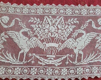 May include: White lace textile with a repeating pattern of floral arrangements and birds. The lace is set against a red background. The design includes a central floral basket flanked by two birds.