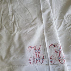 May include: White linen sheet with embroidered initials "M" and "A" in red and blue. The sheet is folded, showing the embroidered details on the lower edge. The fabric has a slightly wrinkled texture.