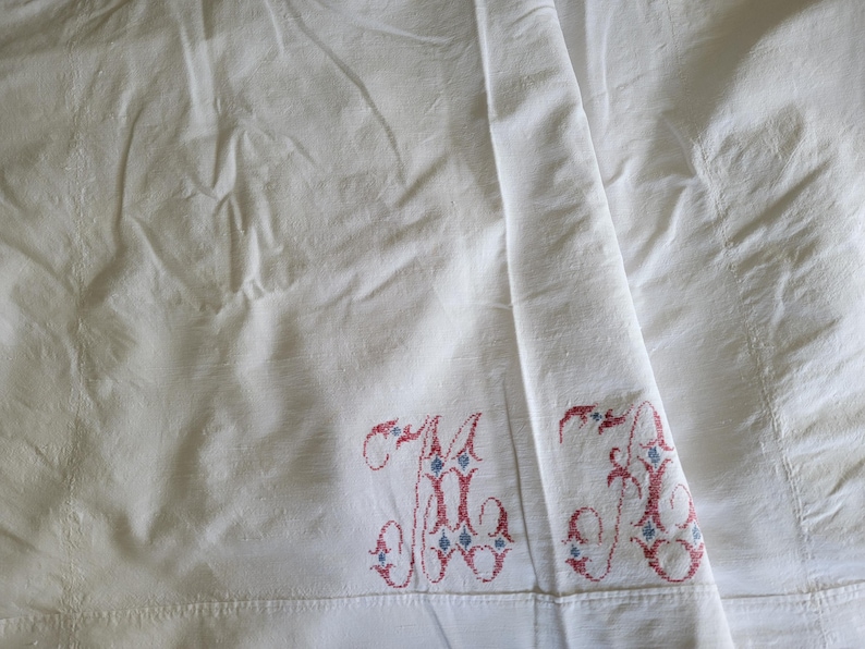 May include: White linen sheet with embroidered initials "M" and "A" in red and blue. The sheet is folded, showing the embroidered details on the lower edge. The fabric has a slightly wrinkled texture.