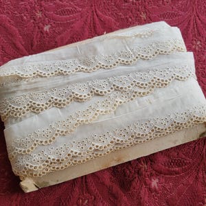 May include: A roll of white cotton lace trim with a scalloped edge and a delicate eyelet pattern. The lace is folded in thirds and has a beige color.