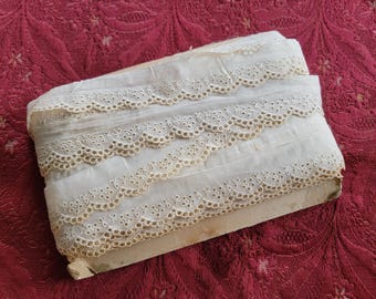 May include: A roll of white cotton lace trim with a scalloped edge and a delicate eyelet pattern. The lace is folded in thirds and has a beige color.