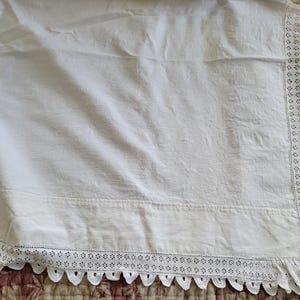 May include: White linen fabric with a decorative lace trim along the edge. The fabric has a slightly textured appearance and is draped over a patterned surface. The lace trim features a scalloped edge.