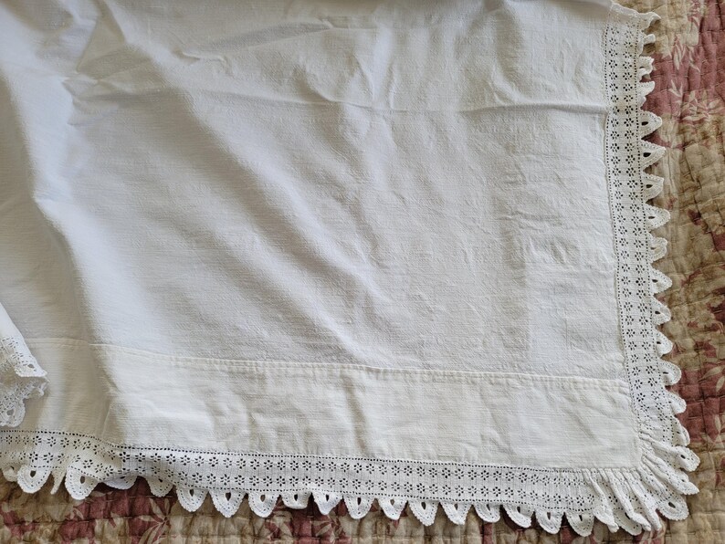 May include: White linen fabric with a decorative lace trim along the edge. The fabric has a slightly textured appearance and is draped over a patterned surface. The lace trim features a scalloped edge.