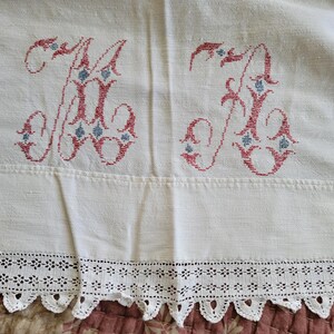 May include: White linen textile with two embroidered "K" monograms in red and blue. The bottom edge features a delicate, scalloped lace trim. The fabric has a slightly textured appearance.