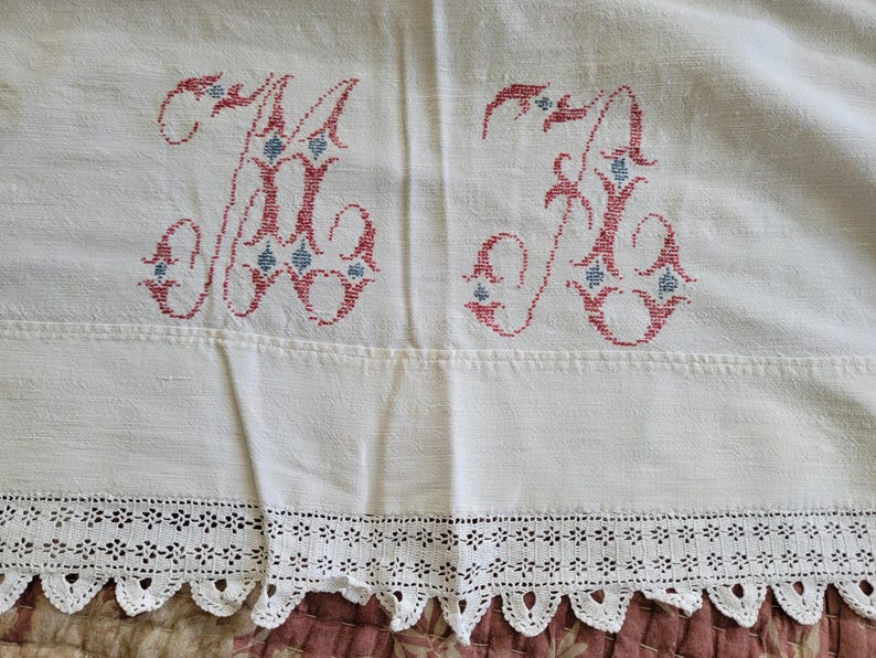 May include: White linen textile with two embroidered "K" monograms in red and blue. The bottom edge features a delicate, scalloped lace trim. The fabric has a slightly textured appearance.