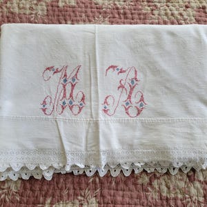 May include: White linen sheet with embroidered initials "M K" in pink and blue. The sheet features a scalloped lace trim along the bottom edge. The sheet is folded and displayed on a floral patterned surface.