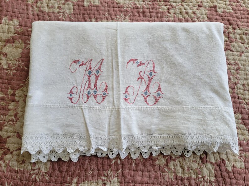 May include: White linen sheet with embroidered initials "M K" in pink and blue. The sheet features a scalloped lace trim along the bottom edge. The sheet is folded and displayed on a floral patterned surface.