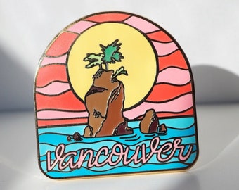 May include: A gold-colored enamel pin with a sunset scene over a small island with palm trees. The text "Vancouver" is written in pink cursive on the blue water.