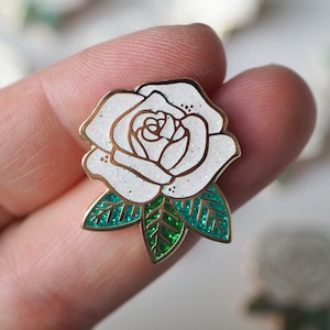 May include: A white rose enamel pin with gold trim and glitter detailing. The pin has green glitter leaves and is held between fingers. Other similar pins are blurred in the background.