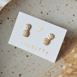 May include: A pair of gold pineapple stud earrings on a white card with the text "Lana Betty".