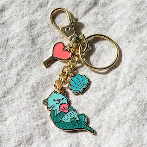 May include: Gold-coloured keychain with a lobster clasp and key ring. The keychain features a teal otter holding a pink shell, a pink heart-shaped key, and a teal seashell charm. The charms are connected by a gold chain.