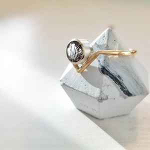 May include: A silver ring with a black and white stone set in a gold band. The ring is displayed on a white and black geometric ring holder.