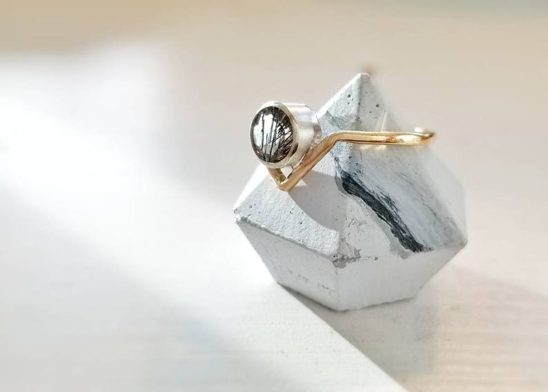 May include: A silver ring with a black and white stone set in a gold band. The ring is displayed on a white and black geometric ring holder.