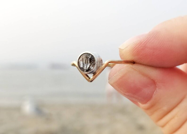 May include: A silver ring with a gold band and a clear gemstone with black inclusions. The ring is shaped like a V.