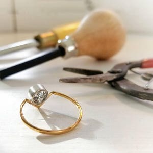 May include: A gold ring with a single round diamond set in a bezel setting. The ring is on a white surface with tools used for jewellery making.