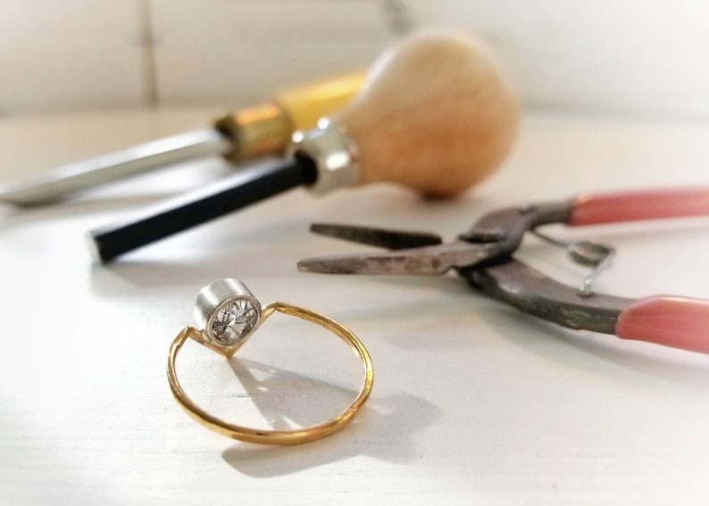 May include: A gold ring with a single round diamond set in a bezel setting. The ring is on a white surface with tools used for jewellery making.