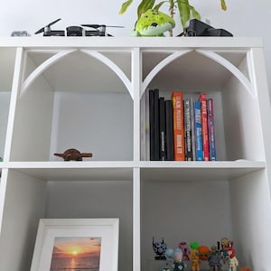 May include: A white four-compartment shelving unit with arched decorative trim. The shelves hold a small wooden turtle, a row of books including titles like Drive and Odine, a framed sunset photograph, and a collection of small colourful figurines.