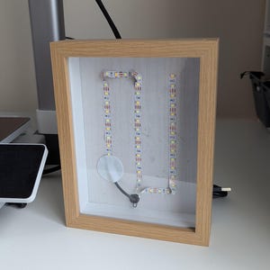 May include: A framed LED light display with a light wood frame. The LED lights are arranged in a vertical pattern with a circular element at the bottom. A black USB cable extends from the frame.