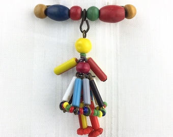 May include: A colourful wooden bead brooch with a dangling figure made of various coloured beads. The figure has a yellow head, red arms, and a skirt made of blue, green, red, and white beads.