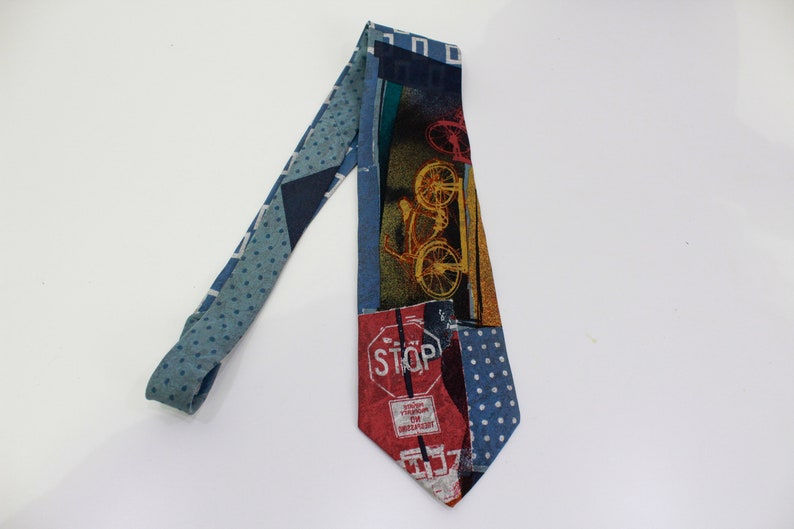 May include: A patterned necktie with a bicycle and stop sign design. The tie features a mix of blue, red, and yellow colors. The tie has a polka dot pattern on the narrow end and a bicycle graphic on the wide end.