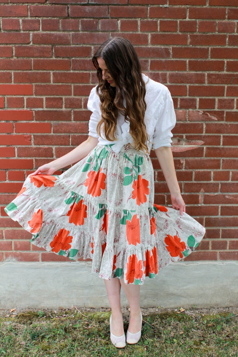 May include: A woman wearing a white blouse tied at the waist and a tiered skirt with a floral print in shades of orange, green, and white. The skirt has a ruffled hem.