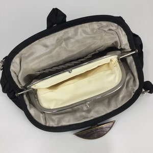Pode incluir: Uma bolsa de m&atilde;o de tecido preto com um fecho de prata e um detalhe em metal castanho. A bolsa est&aacute; aberta e o interior &eacute; forrado com um tecido bege.