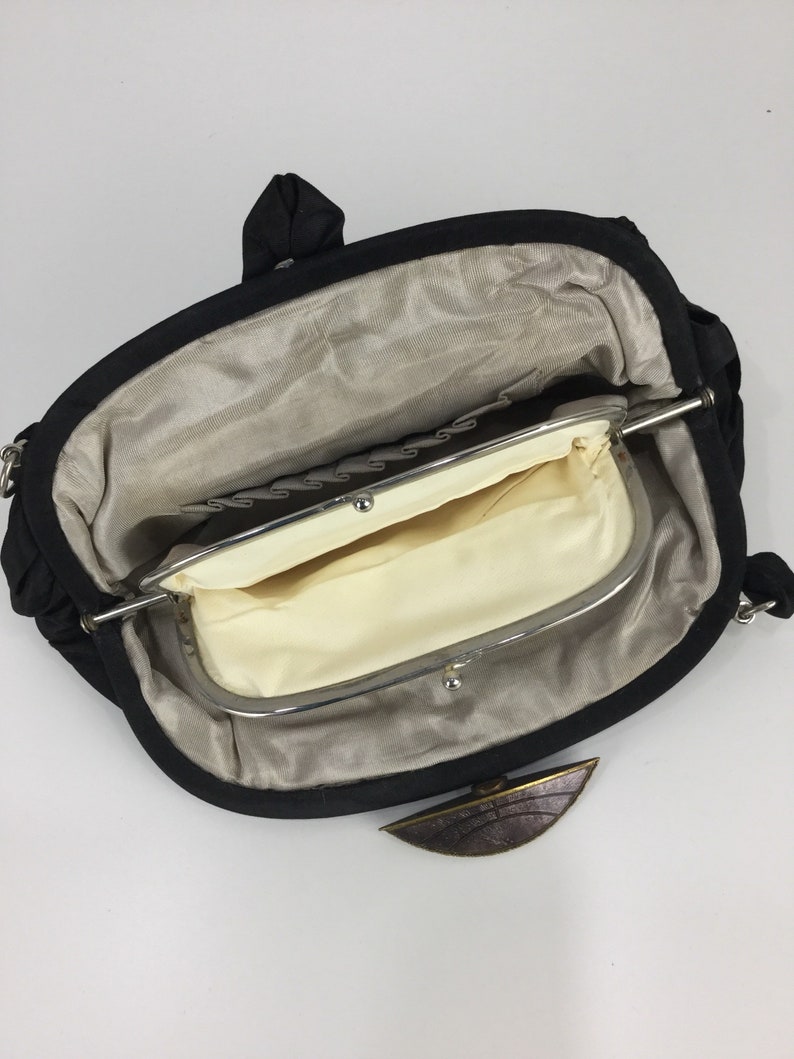 Pode incluir: Uma bolsa de m&atilde;o de tecido preto com um fecho de prata e um detalhe em metal castanho. A bolsa est&aacute; aberta e o interior &eacute; forrado com um tecido bege.