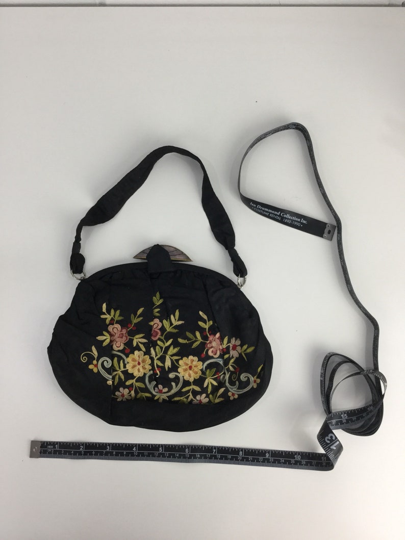 Pode incluir: Uma bolsa de m&atilde;o de cetim preto bordada com um design floral e um fecho de madeira. A bolsa tem uma al&ccedil;a comprida e mede aproximadamente 18 cm de largura.
