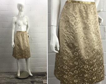 May include: A gold-colored skirt with a floral pattern. The skirt is knee-length and has a waistband with a gold trim.