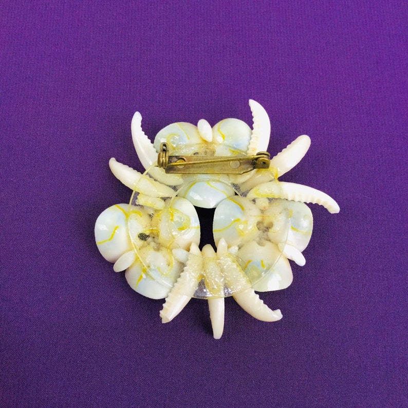 May include: A white and yellow seashell brooch with a gold pin. The brooch is made of multiple seashells arranged in a circular pattern.