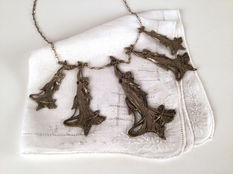 May include: A silver necklace with five pendant charms, each featuring a stylised female figure with flowing drapery. The necklace is displayed on a white fabric background.