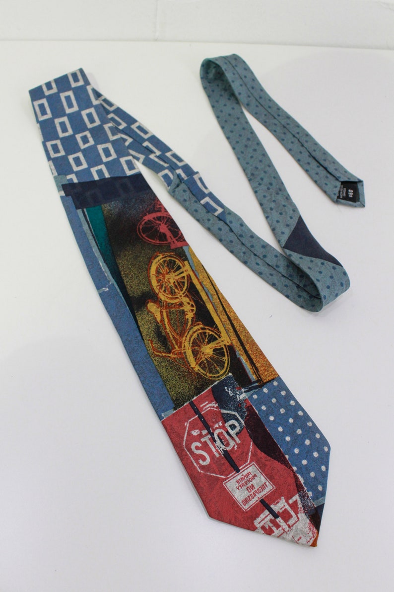 May include: A patterned necktie with a patchwork design. The tie features various patterns, including blue squares, polka dots, and a bicycle graphic. The bottom of the tie has a red stop sign graphic. The tie is made of a soft fabric.