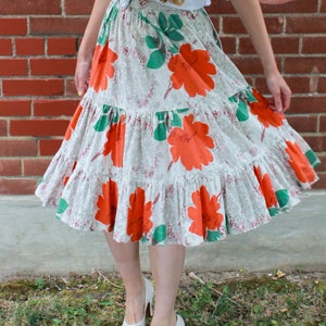 May include: A white skirt with a floral print featuring large orange flowers and green leaves. The skirt has multiple tiers and is gathered at the waist.