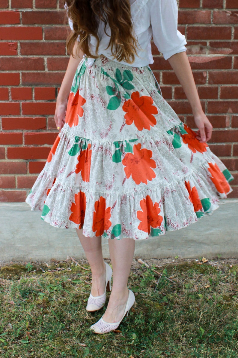 May include: A white skirt with a floral print featuring large orange flowers and green leaves. The skirt has multiple tiers and is gathered at the waist.