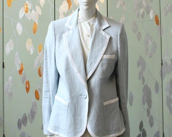 Pode incluir: Blazer azul claro com acabamento branco, usado sobre uma blusa e saia creme. O blazer tem um fecho de um botão e dois bolsos frontais. O conjunto é exibido em um manequim.