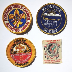 May include: Four embroidered patches in yellow, red, blue and white. The patches are circular and feature text such as "Pokagon Trails Council", "Jubilee for God and Country", "Klondike Ashkum District 1961 Derby", and "Camp Betz Pokagon Trails Council Boy Scouts of America".