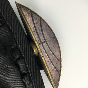 Pode incluir: Um close-up de um fecho met&aacute;lico decorativo com um design circular texturizado. O fecho &eacute; de cor bronze escuro com um padr&atilde;o prateado.
