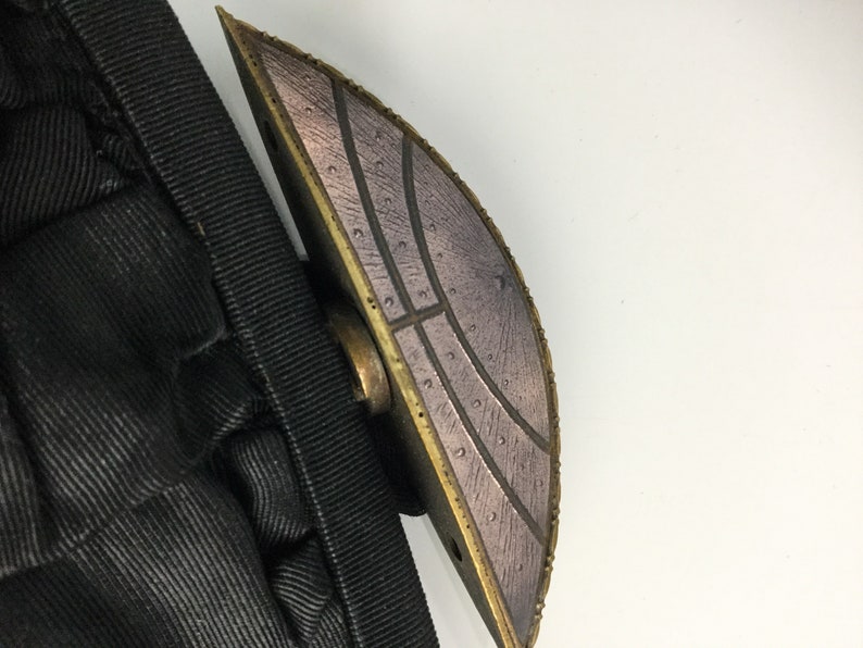 Pode incluir: Um close-up de um fecho met&aacute;lico decorativo com um design circular texturizado. O fecho &eacute; de cor bronze escuro com um padr&atilde;o prateado.