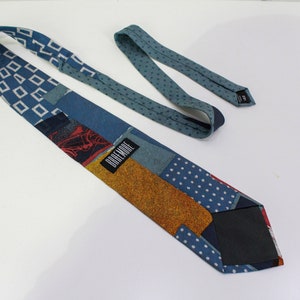 May include: A patchwork necktie with various patterns and colors. The tie features blue geometric shapes, polka dots, and a textured gold section. The label reads "DREMORE".