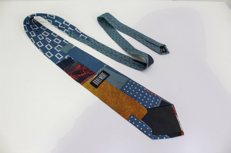May include: A patchwork necktie with various patterns and colors. The tie features blue geometric shapes, polka dots, and a textured gold section. The label reads "DREMORE".