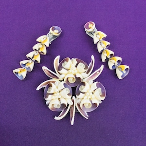 May include: A set of three hair clips made from seashells. The clips are in the shape of flowers with white and purple shells. The clips are on a purple background.