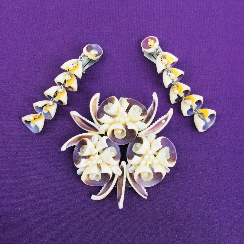 May include: A set of three hair clips made from seashells. The clips are in the shape of flowers with white and purple shells. The clips are on a purple background.