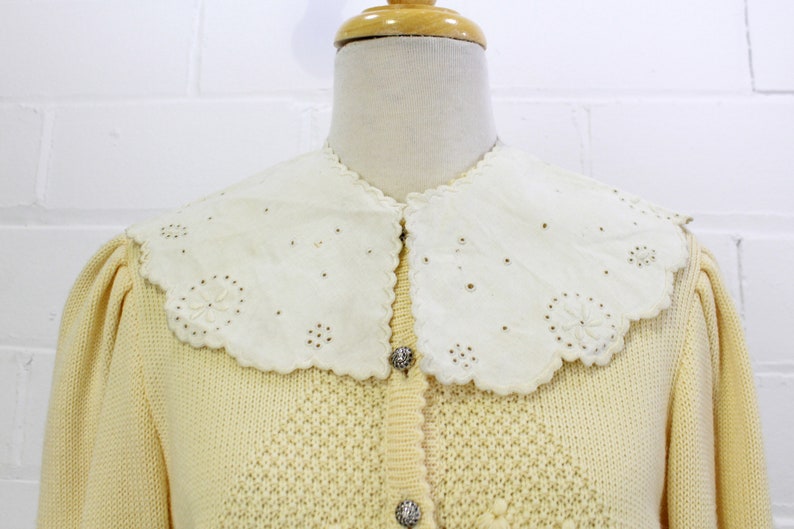May include: A pale yellow knitted cardigan with a white crocheted collar and pearl-like buttons.