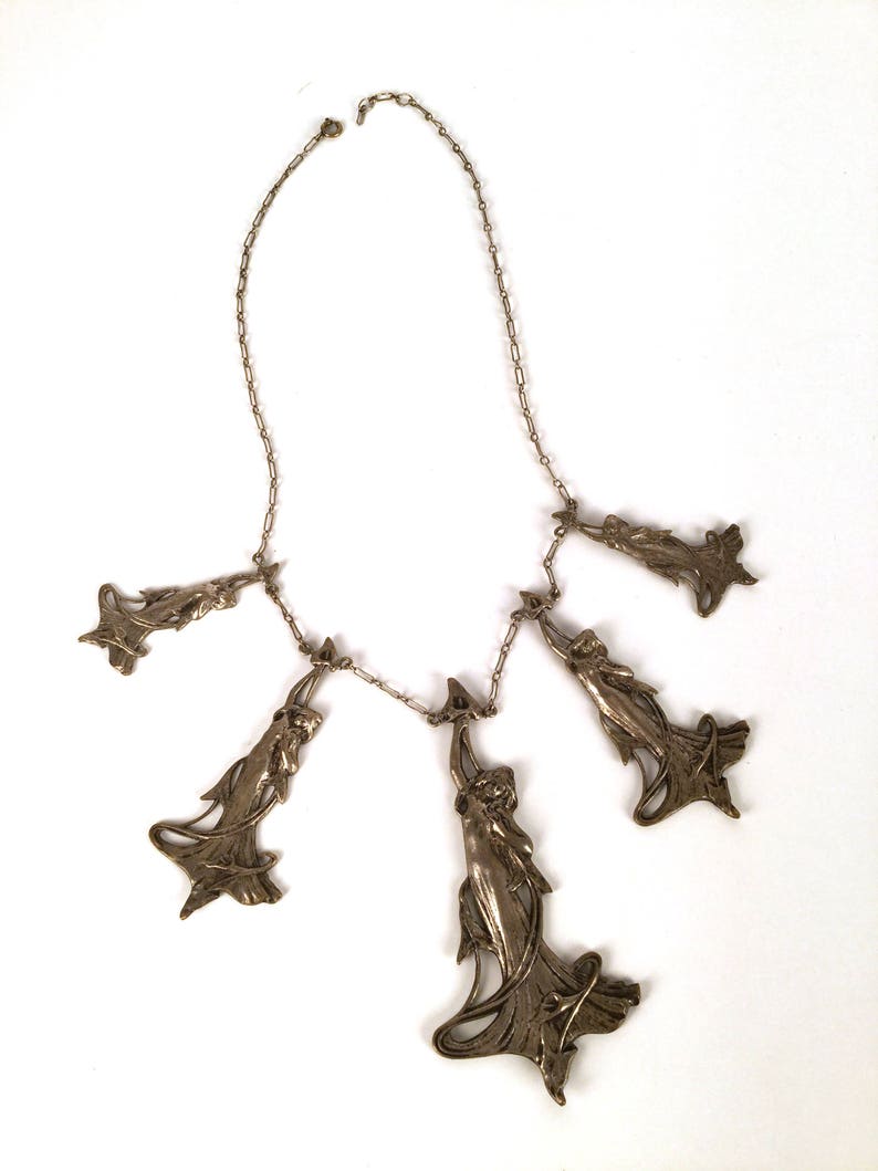 May include: A silver necklace with five pendant charms, each featuring a stylised female figure in a flowing gown. The charms are connected to a delicate silver chain.
