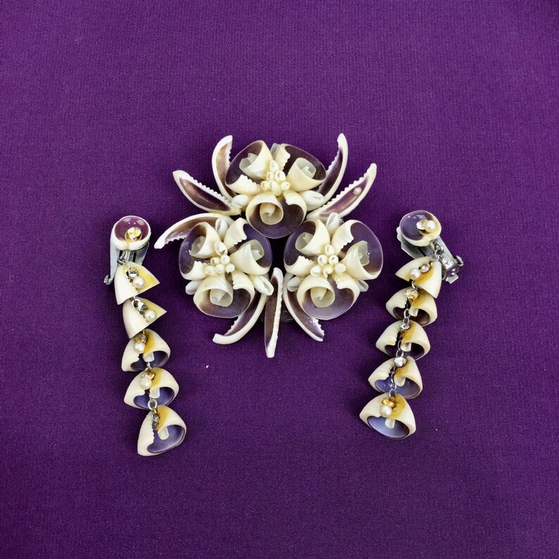 May include: A set of shell jewelry featuring a brooch and a pair of earrings. The brooch is a cluster of white and purple seashells arranged in a flower shape. The earrings are long and dangle, with each earring made of a chain of white and purple seashells.