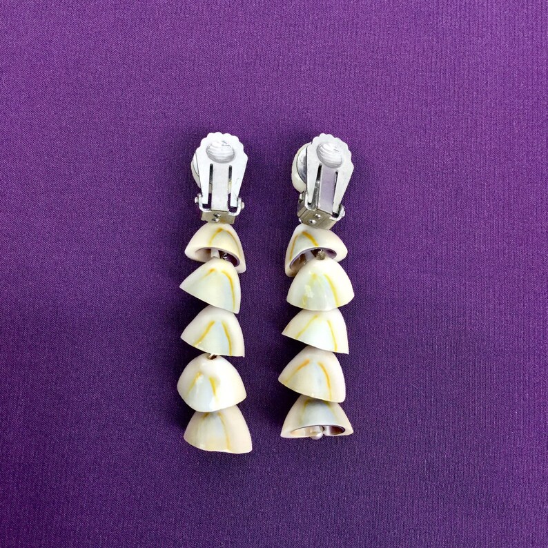 May include: A pair of clip-on earrings made of white seashells with a yellow and brown pattern. The earrings are made of five seashells each, and the clip-on backs are silver.
