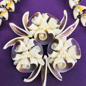 May include: A close-up of a floral brooch made from seashells. The brooch is made of white and purple seashells arranged in a flower shape. The brooch is a beautiful and unique piece of jewelry.