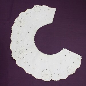 May include: White cotton lace collar with a scalloped edge and delicate floral embroidery. The collar is designed to be worn around the neck and has a rounded shape.