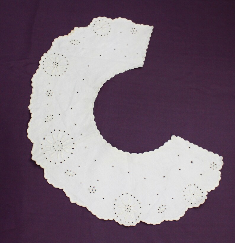 May include: White cotton lace collar with a scalloped edge and delicate floral embroidery. The collar is designed to be worn around the neck and has a rounded shape.